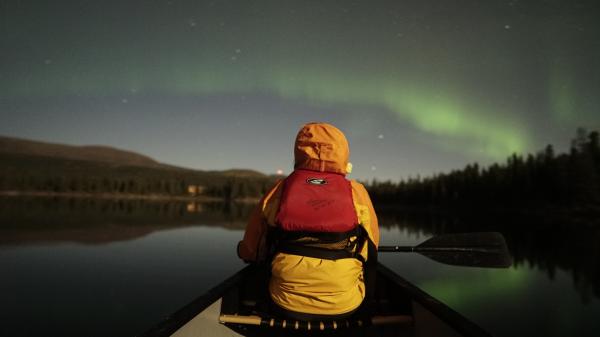 A person in a yellow jacket and red life jacket watches the aurora borealis from the front of a canoe