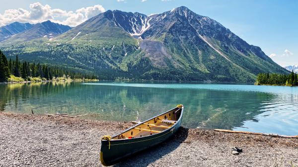 A canoe is on the shore of a clear teal lake with a large mountain in the background