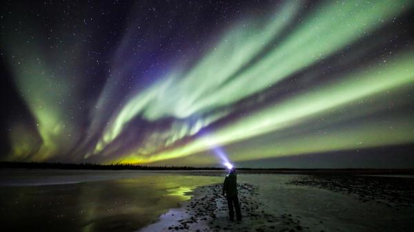 Aurora shines over someone in front of a lake wearing a headlamp