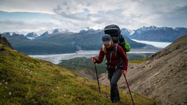 A woman is hiking up a green slope with a large backpack and hiking poles. 