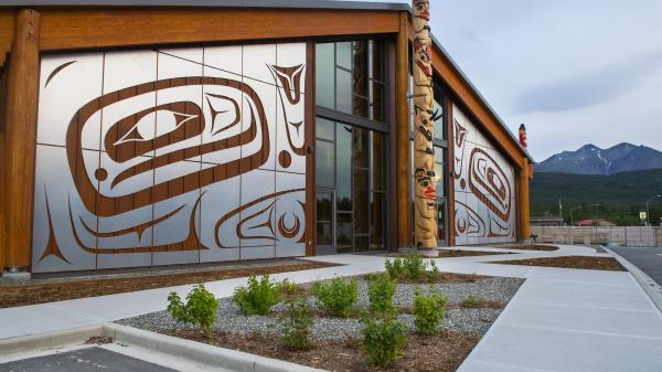 A building adorned with Carcross Tagish First Nations artwork sits under a flat grey sky. A totem pole stands right outside the doors.