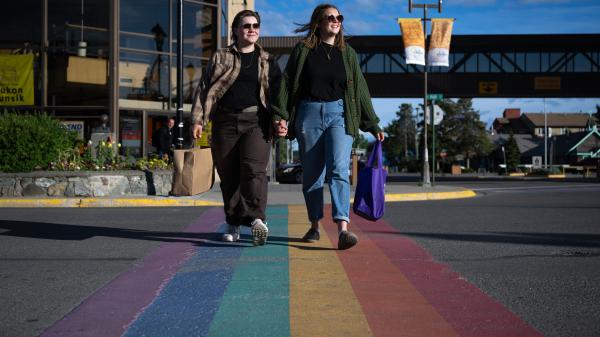 A queer couple steps over a rainbow crosswalk