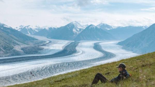 A man sits on a grassy field overlooking a mountain and glacier