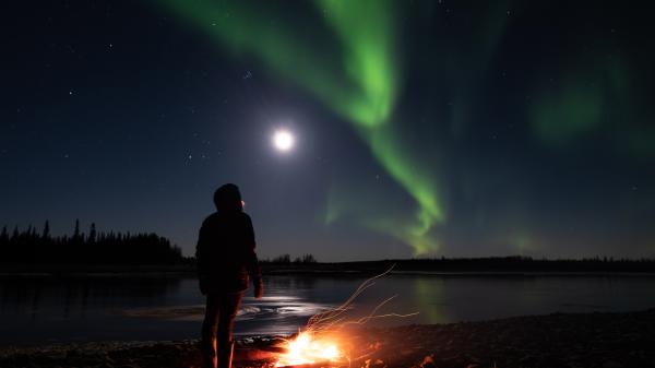 A person stays warm by a campfire while they watches the aurora borealis.