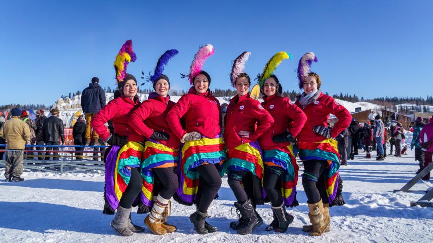 A group of can-can dancers wearing matching colourful skirts and red jackets stand in a line. They each have a feather in their hair.
