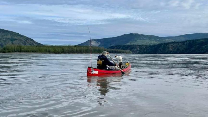 A red canoe with a person and a dog. Forest and mountains in background