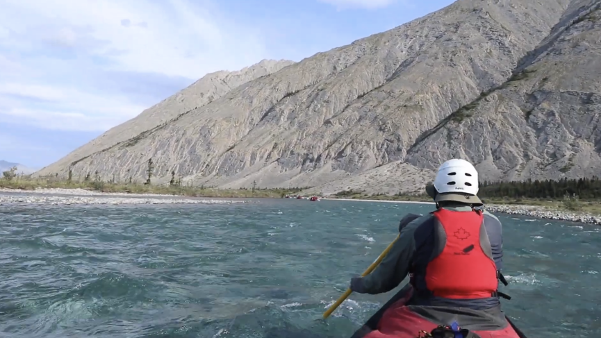 A bow paddler takes a stroke on the left as the canoe float through gentle, clear water with a small amount of current, with light grey and brown treeless mountains in the background.