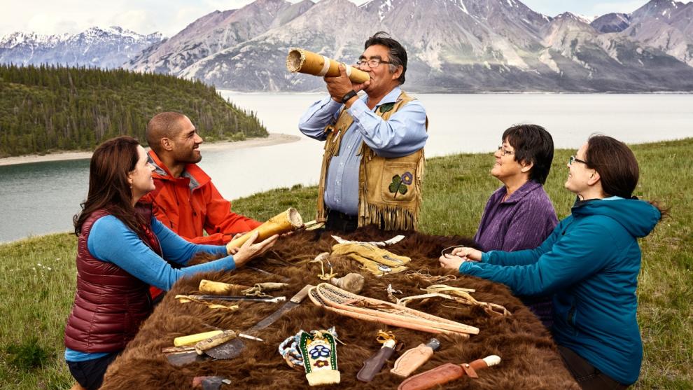 A first nations man blows into a horn in front of people seated around a picnic table