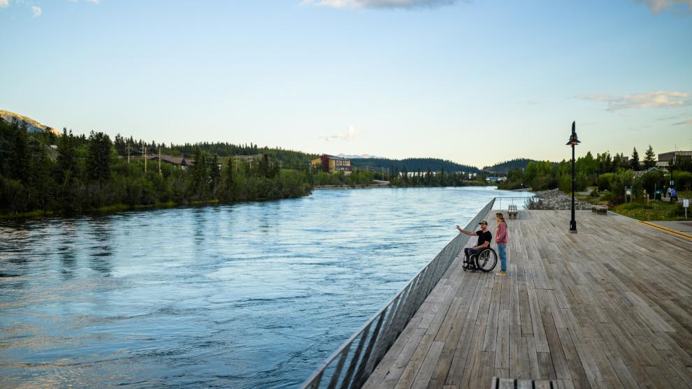 A man in a wheelchair chats with a friend on the accessible viewing platform along the Yukon River (Tágà Shäw) waterfront in downtown Whitehorse.