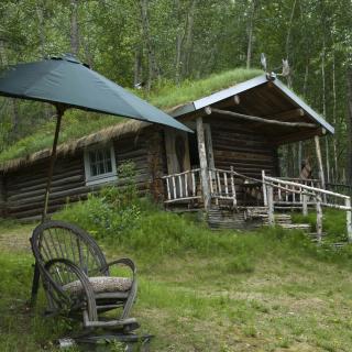 A small cabin sits on a green hillside behind a wicker chair and a large umbrella
