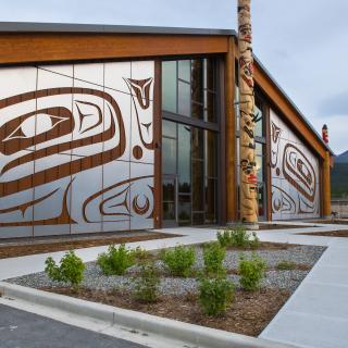 A building adorned with Carcross Tagish First Nations artwork sits under a flat grey sky. A totem pole stands right outside the doors. 
