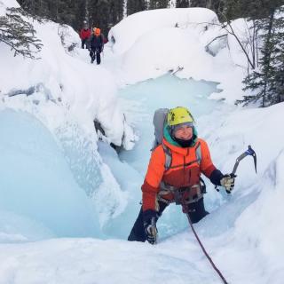 Woman in bright orange jacket smiling while ice climbing frozen waterfall