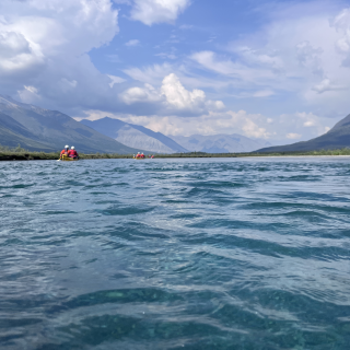 Canoes float down aquamarine water in a mountainous river valley with the sun shining.
