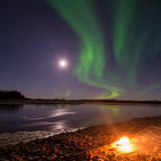 Northern lights swirl above open water and a campfire