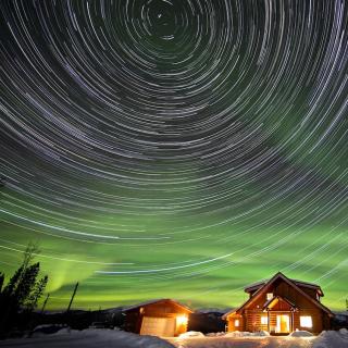 stars swirl among aurora above a cabin glowing in the night