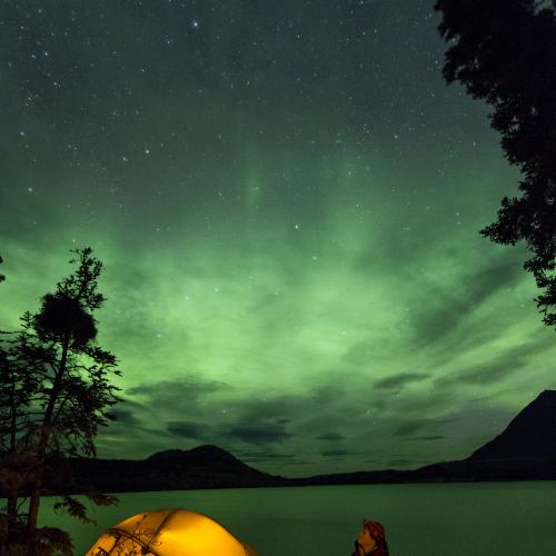 Someone sitting outside of a tent near a lake watches the northern lights