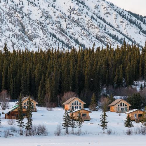 Cozy wooden cabins nestled in snowy Yukon forest with towering mountain backdrop.