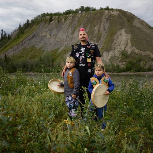 Derrick standing with two children who are holding drums against background of a mountain and a river. 