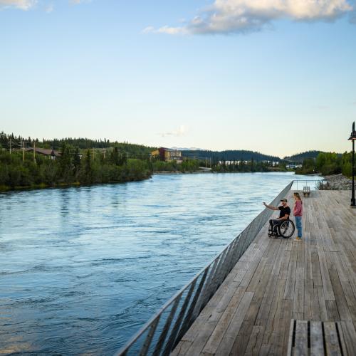 A man in a wheelchair chats with a friend on the accessible viewing platform along the Yukon River (Tágà Shäw) waterfront in downtown Whitehorse.