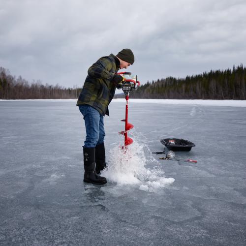Gavin Winter Sinnott drills a fishing hole through lake ice with an auger on a frozen lake.