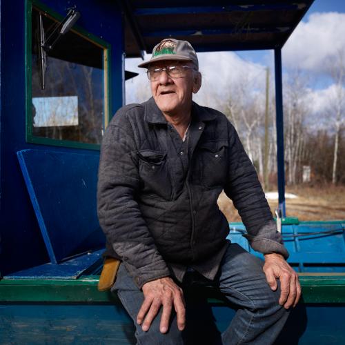 Don Trudeau wearing a baseball cap and glasses seated on the edge of a bright blue wooden structure outdoors.