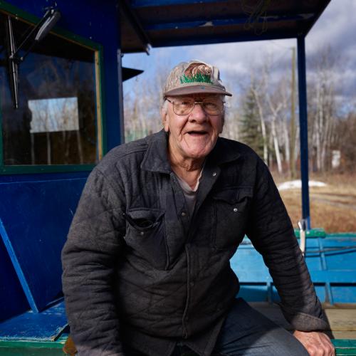 Don Trudeau wearing a baseball cap and glasses seated on the edge of a bright blue wooden structure outdoors.