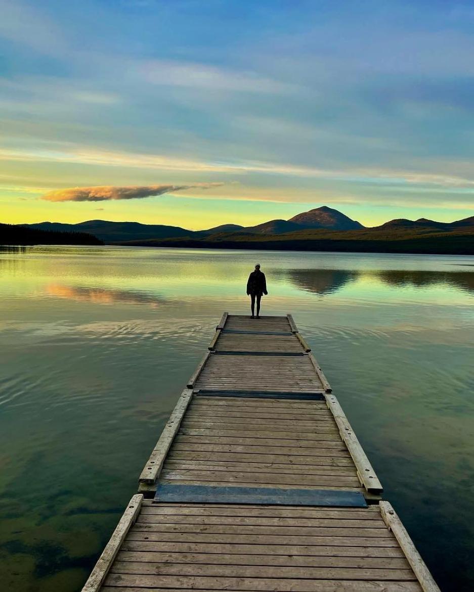A person standing on the end of a long dock overlooks a large lake