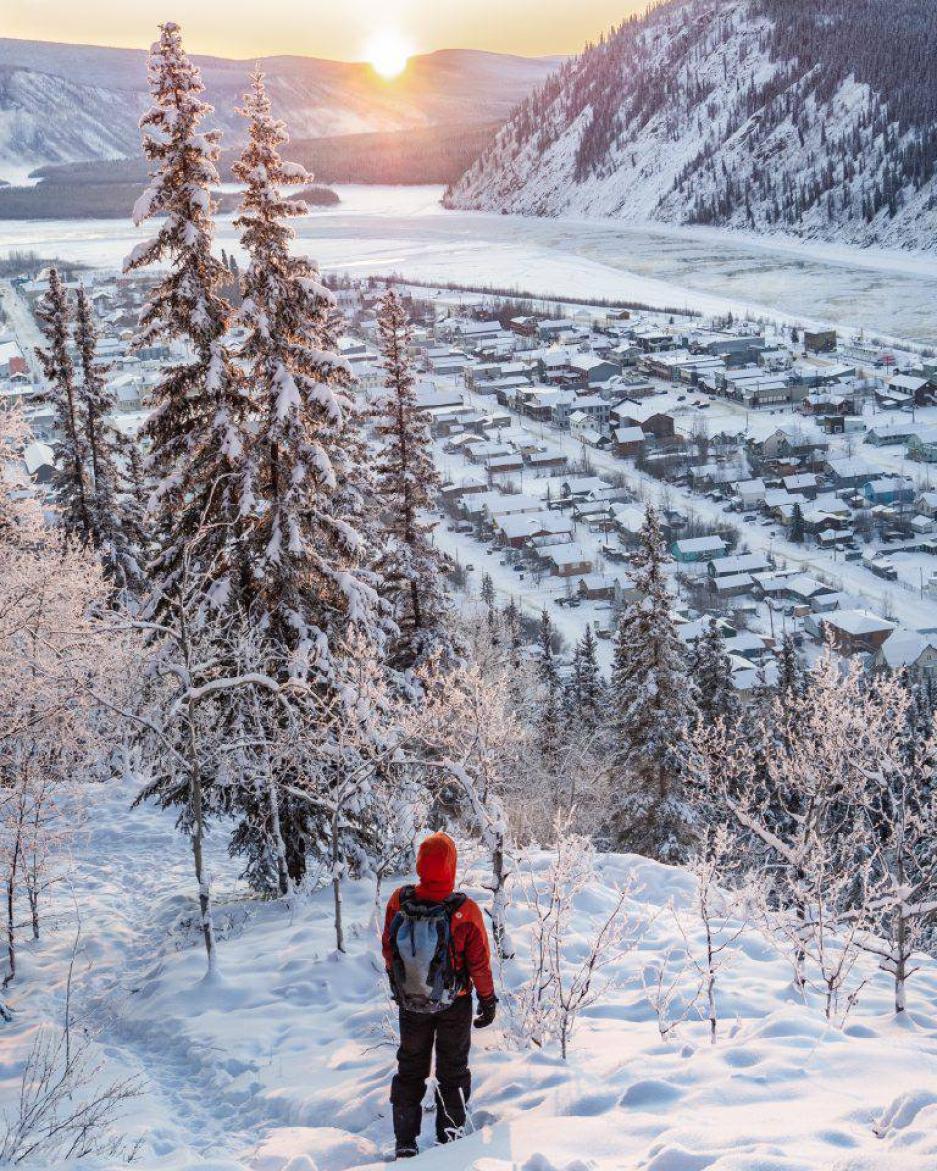 A hiker in a red jacket makes their way down a snowy mountain toward Dawson City
