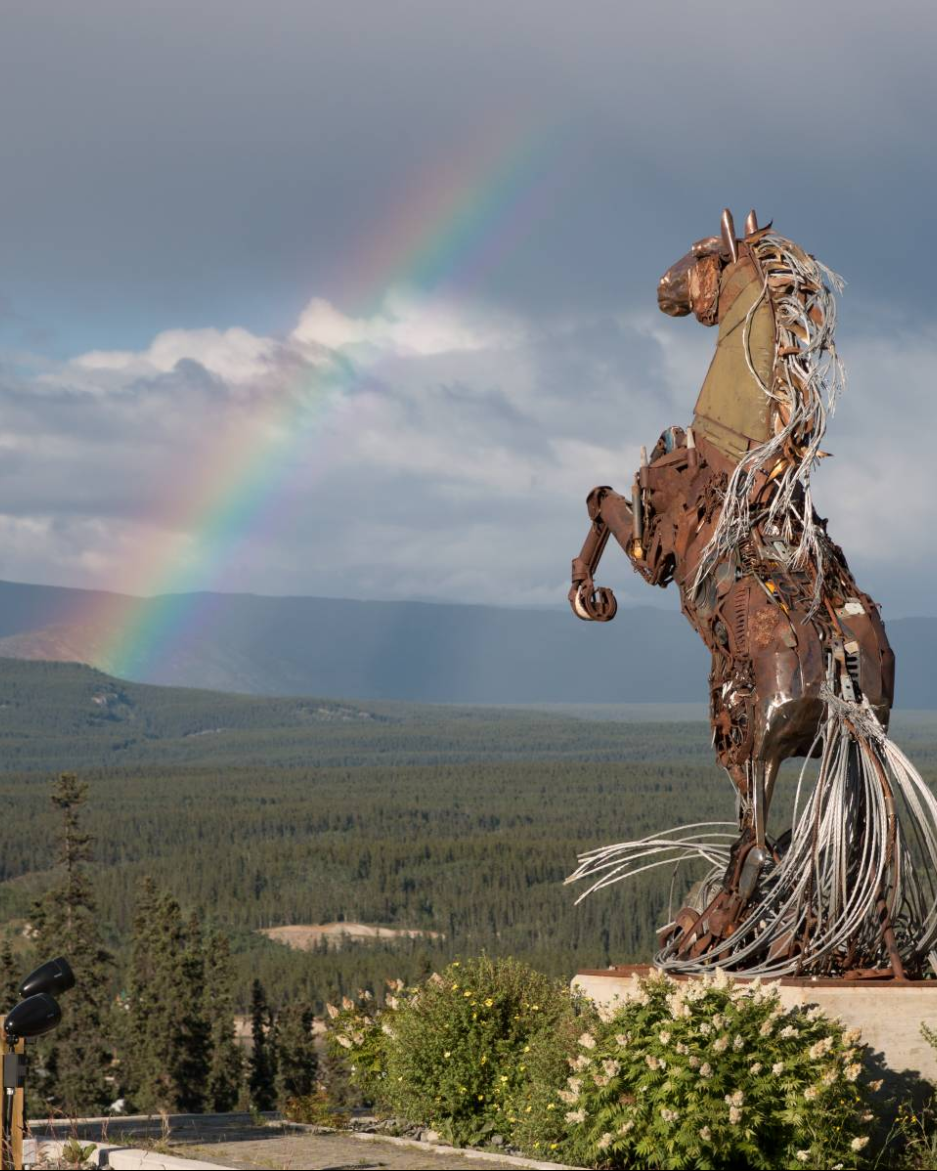 A metal horse structure stands tall over a mountain valley with a rainbow shining over it