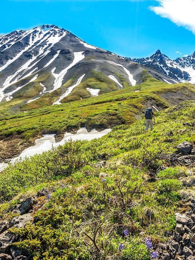 A man hikes up a green valley-side towards a mountain with the reminants of snow