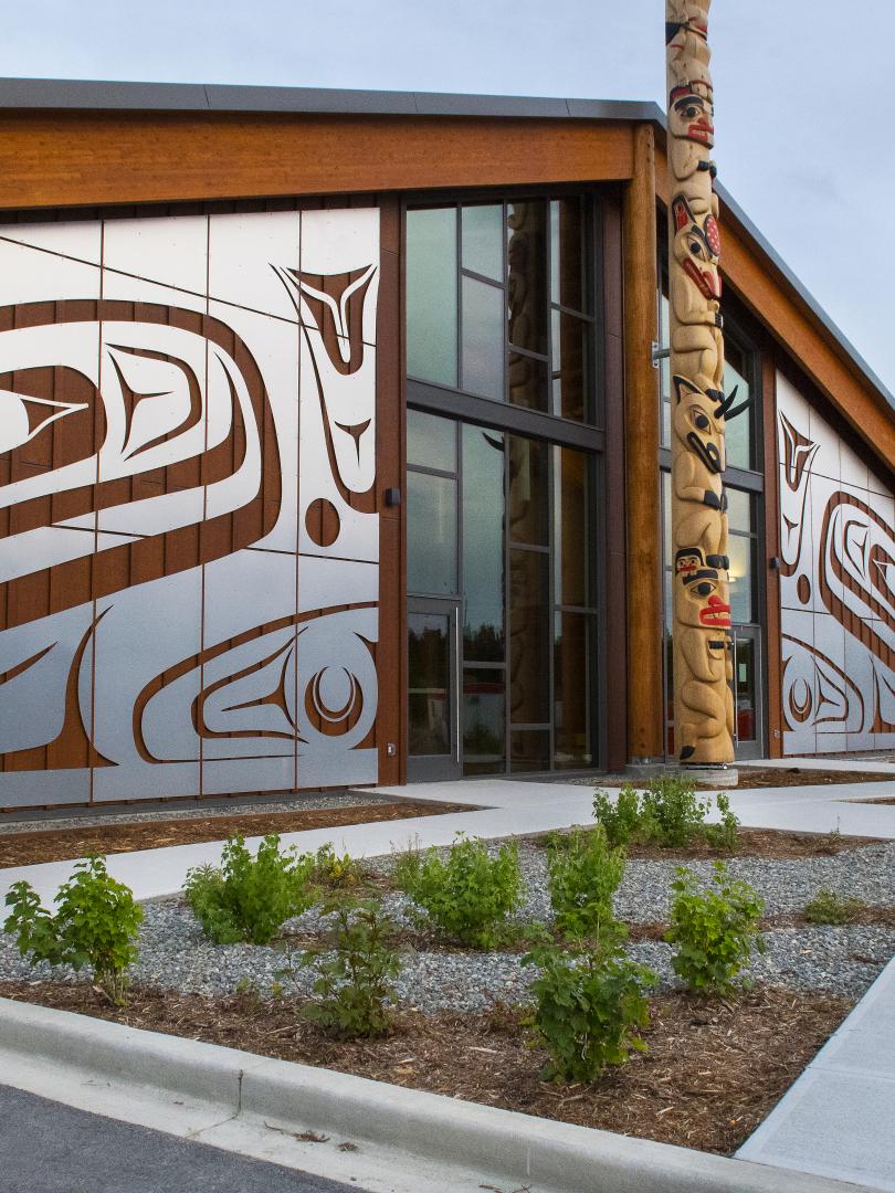 A building adorned with Carcross Tagish First Nations artwork sits under a flat grey sky. A totem pole stands right outside the doors. 