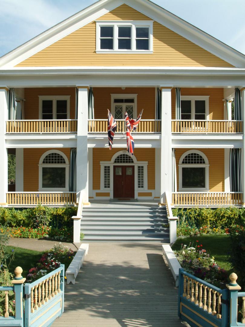 A yellow house with a white picket fence, green front lawn and tall white pillars