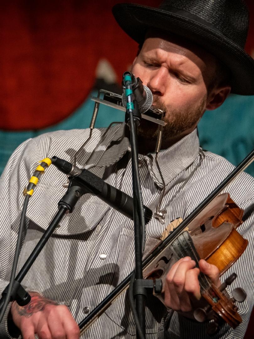 A man in a black hat plays the violin and sings longingly into a microphone