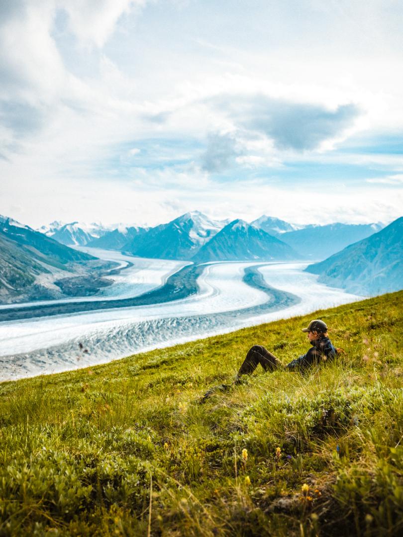 A hiker takes a break, lying down and admiring the breathtaking view of a glacier in Kluane National Park, Yukon. Experience the stunning landscapes and serene beauty of this wild and remote Canadian wilderness on an unforgettable guided hiking adventure.