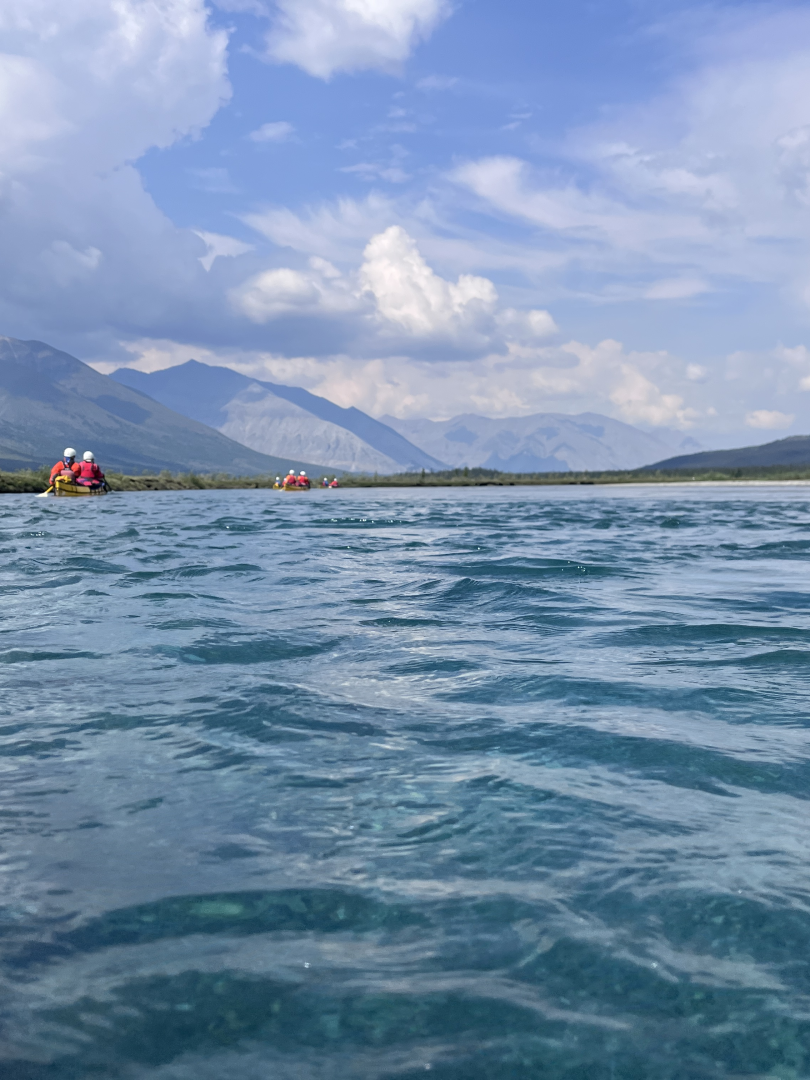 Canoes float down aquamarine water in a mountainous river valley with the sun shining.