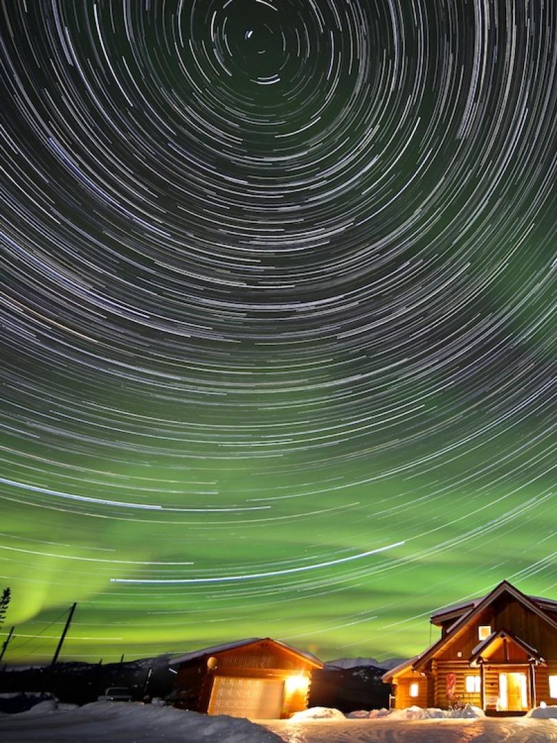 stars swirl among aurora above a cabin glowing in the night