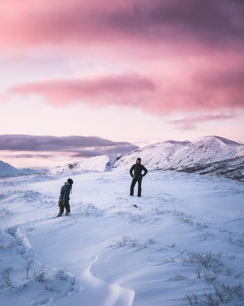 Two hikers walk across a snowy mountain plateau under a pink sky