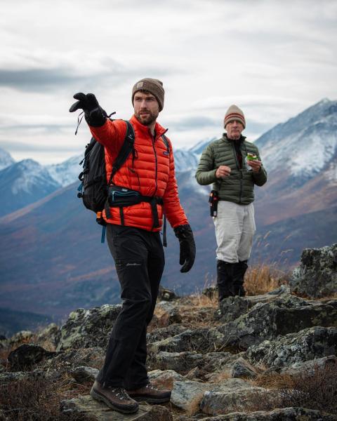 Two men on a mountain top stand and point at something in the distance. Snowcapped mountains are in the background.