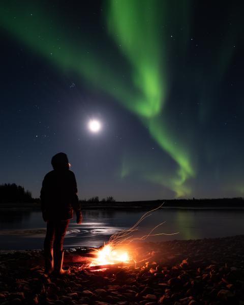 A person stays warm by a campfire while they watches the aurora borealis. 
