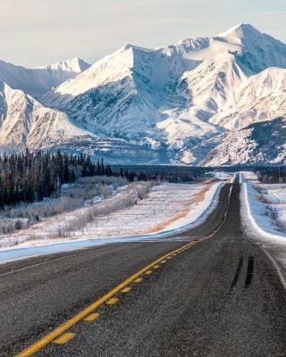 A paved road with yellow lines stretches toward a tall, snow covered mountain