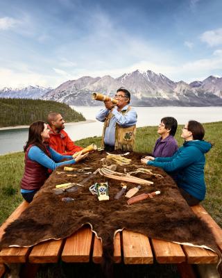 A first nations man blows into a horn in front of people seated around a picnic table