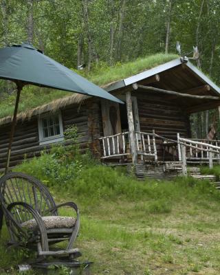 A small cabin sits on a green hillside behind a wicker chair and a large umbrella