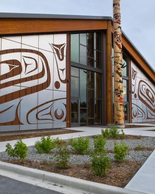 A building adorned with Carcross Tagish First Nations artwork sits under a flat grey sky. A totem pole stands right outside the doors. 