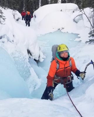 Woman in bright orange jacket smiling while ice climbing frozen waterfall 