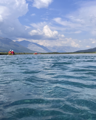 Canoes float down aquamarine water in a mountainous river valley with the sun shining.