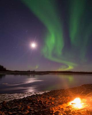 Northern lights swirl above open water and a campfire