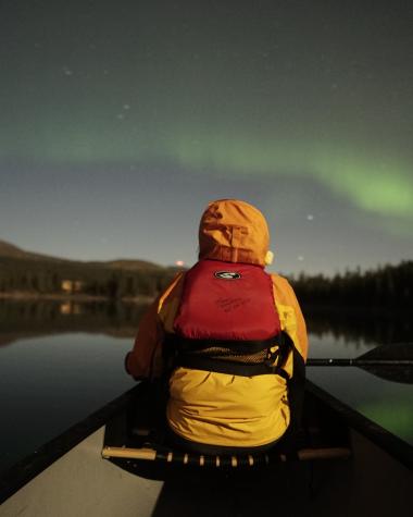 A person in a yellow jacket and red life jacket watches the aurora borealis from the front of a canoe