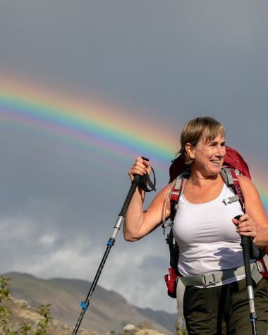 A woman holds two hiking poles and looks off to the side as a rainbow shines over head