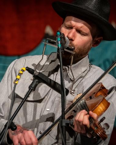 A man in a black hat plays the violin and sings longingly into a microphone