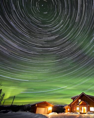 stars swirl among aurora above a cabin glowing in the night